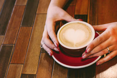 Coffee cappuccino with foam heart in woman hands with beautiful manicure on wooden background