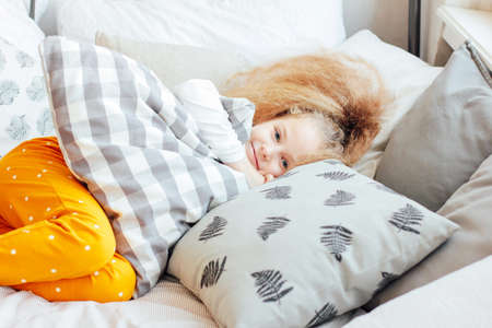 Little beautiful curly blonde girl woke up and smiling, cozy morningの写真素材