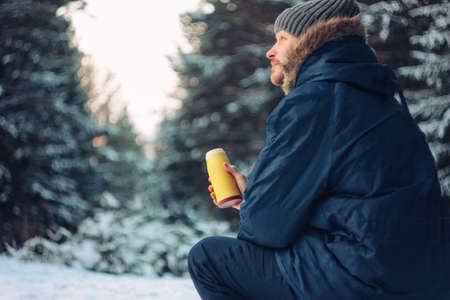 Man traveler forester hunter in the winter forest sitting on fallen tree with flask in his hands