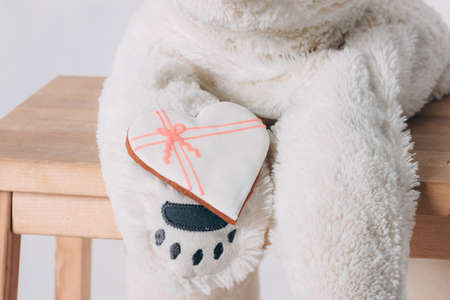White heart gingerbread on paw of old bear toy on wooden chair isolated on grey backgroundの写真素材
