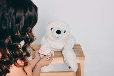 Woman hands and white heart gingerbread on paw of old bear toy on wooden chair isolated on grey...