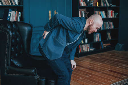The attractive bald bearded man is holding onto his back, trying to get up from his leather chair. Rich people cry tooの写真素材