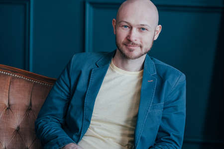 Portrait of attractive adult successful bald bearded man in suit on rich interior blue background