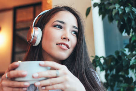 Beautiful charming brunette smiling Asian girl enjoys music in headphones with cup of coffee or tea...