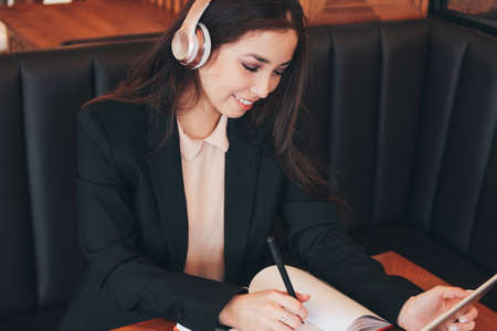 Beautiful charming brunette smiling asian girl in headphones with tablet in cafe, student, freelancer. Learning foreign languages or online coursesの写真素材