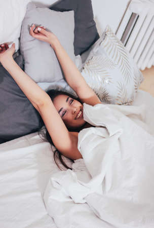 Happy smiling beautiful long hair asian girl young woman lying down in bed, cozy morningの写真素材