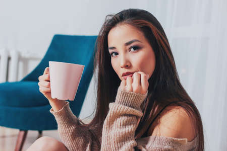 Candid portrait of sensual smiling asian girl young woman with dark long hair in cozy beige sweater holding cup of tea on white curtain backgroundの写真素材
