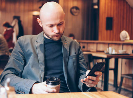 Adult bald gloomy man drinking coffee from paper cup and using mobile phone at cafeの写真素材