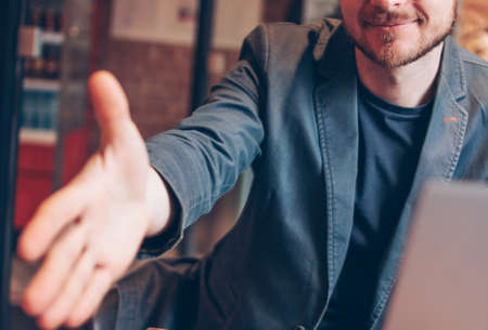 Smiling attractive adult successful bald bearded man in suit with laptop giving handshake, hand of help, greeting at cafeの写真素材