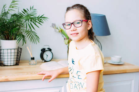 Pretty smiling little girl in glasses and pigtails sitting with book at the table in her roomの写真素材