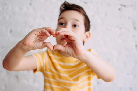 Happy tweens boy in yellow t-shirt making heart by hands on the white wall background, selective focusの写真素材