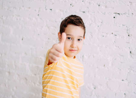 Portrait of cute tween smiling boy in yellow t-shirt shows thumb up isolated on white brick wall backgroundの写真素材