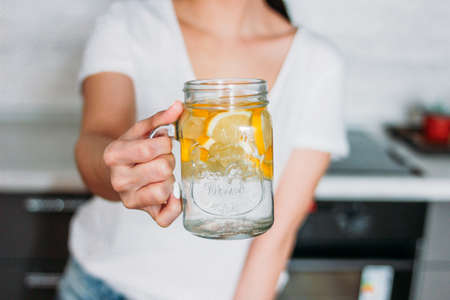 The slim girl young woman holding glass jar with lemon water in kitchen, close up