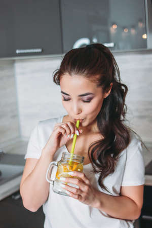 The beautiful asian girl young woman drinking lemon water from glass jar in kitchenの写真素材