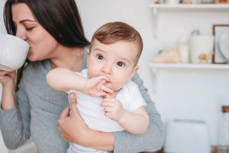 Young mother brunette woman with baby boy in arms drinking tea in white modern kitchen at homeの写真素材