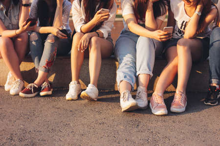 Crop photo of group of young happy tan girls real friends using mobile on sunset backgroundの写真素材