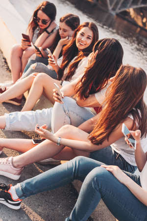 Group of young happy girls real friends students using mobile at city street on sunset backgroundの写真素材