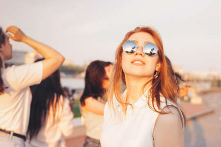 Positive beautiful happy red haired girl in mirror sunglasses with friends on blue sky background, summer sunset timeの写真素材