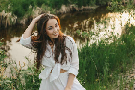 Beautiful carefree long hair asian girl in the white clothes on river side in the grass at sunset. Sensitivity to nature conceptの写真素材