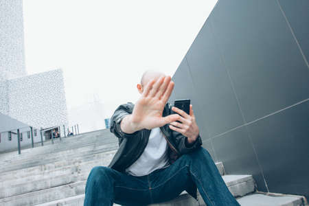 Adult bald bearded serious man with mobile sitting in the stair closing camera by hand, no more...