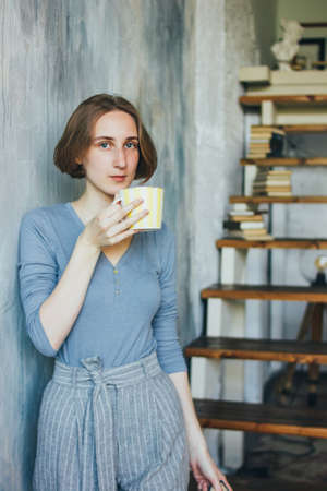 Young woman in with yellow cup standing against wall in the art work studio, vertical portrait
