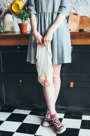 Young woman in grey dress with knitted rag bag string bag shopper in the kitchen, zero waste, slow...