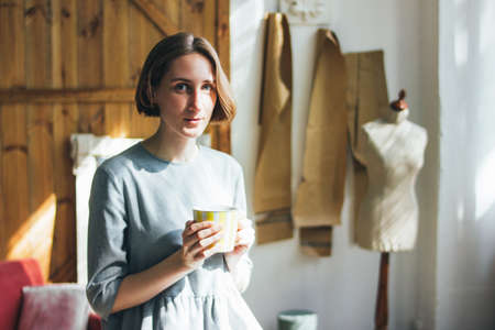 Young woman in simple grey dress with yellow cup sitting on chair in art work studio, full-length...