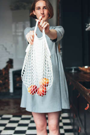 Young woman in the grey dress pulls apples out of knitted rag bag string bag shopper in the kitchen,...