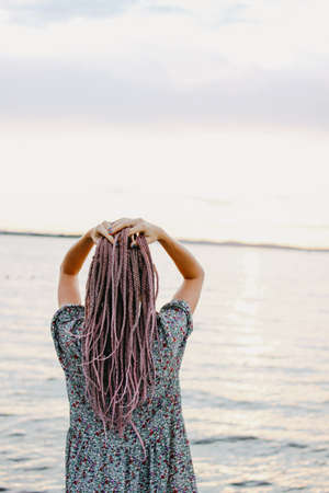 Beautiful romantic young woman with african braids in dress on beach, view from behind, summer vacation timeの写真素材