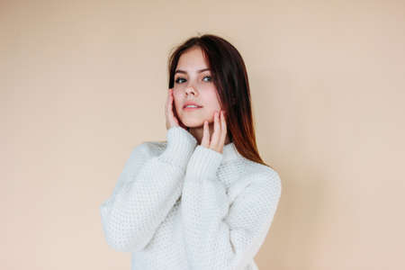 Beautiful girl with clean skin and dark long hair in cozy white sweater on the beige backgroundの写真素材