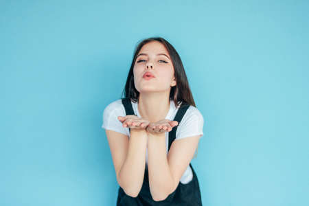 Beautiful carefree smiling girl with dark long hair in white t-shirt making air kiss isolated on the blue backgroundの写真素材