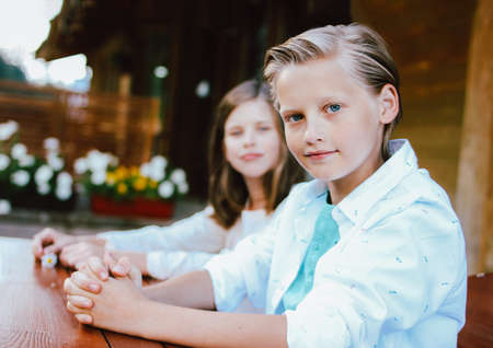 Attractive preteen blonde boy with blue eyes in white shirt with sister friend sitting at the table in backyard of wooden cottageの写真素材