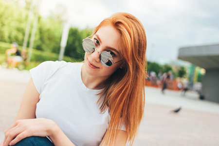 Attractive redhead smiling girl in round sunglasses in casual clothes sitting on street in the cityの写真素材