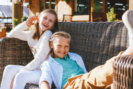 Attractive preteen children sister and brother, friends sitting on wicker sofa in backyard of the wooden cottageの写真素材