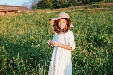 Beautiful romantic preteen girl in white dress and straw hat at the meadow, golden hourの写真素材