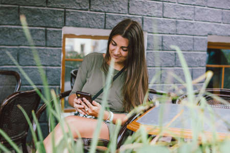 Young smiling long hair brunette girl dressed casual using mobile in street cafeの写真素材