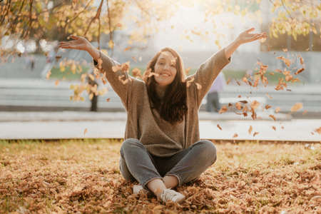 Friendly charming brunette woman with long curly hair sitting on the ground and throws golden leaves, autumn seasonの写真素材