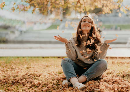 Friendly charming brunette woman with long curly hair sitting on the ground and throws golden leaves, autumn seasonの写真素材