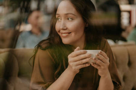 Charming brunette woman with long curly hair sitting at window in cafe with cup of coffee in handsの写真素材