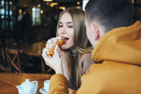 Happy young couple dressed warm casual clothing sitting at cafe together. The man feeds girl a croissantの写真素材