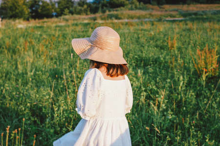 Beautiful romantic preteen girl in white dress and straw hat from behind at the meadow, golden hour