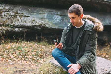 Young male traveler in khaki parka jacket with backpack looks at the route in his mobile phone, travel adventure lifestyleの写真素材