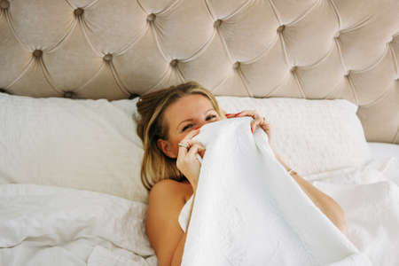 Happy smiling beautiful long fair hair woman in underwear closing face sitting on bed in the bright...