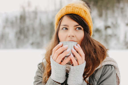 Brunette girl in yellow knitted hat with metal mug of hot tea in the forest outdoors in winterの写真素材