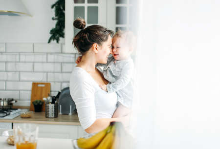 Young woman mom with baby girl on hands havinh fun and cooking breakfast on bright kitchen at the...
