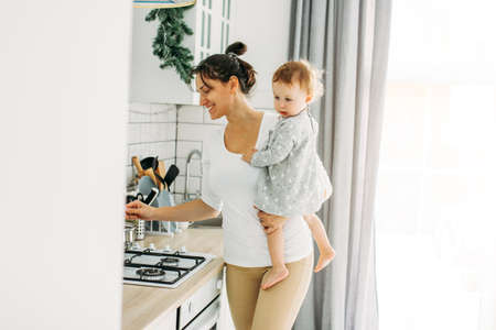 Young woman mom with baby girl on hands cooking breakfast on bright kitchen at the homeの写真素材