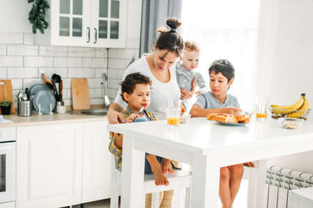 Young woman mom with baby girl on hands cooking breakfast on bright kitchen at home large happy familyの写真素材