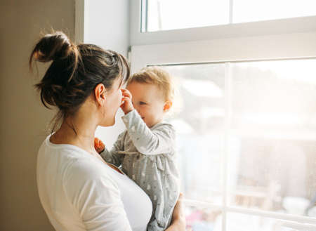 Young woman mom with baby girl on hands near window at the homeの写真素材