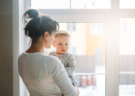 Young woman mom with baby girl on hands near window at the home