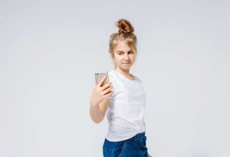 Carefree smiling preteen girl with fair hair in white t-shirt and jeans making selfie, isolated on...